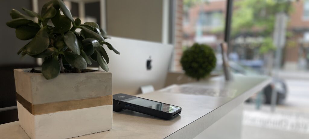 A smartphone and a potted plant rest on a white counter in front of a desktop computer displaying an outdoor scene, with the billing software window subtly open, reflecting the end-of-month fees.